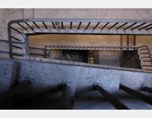 Blick in das Treppenhaus eines Gebäudes mit Betonwänden und Metallgeländern, das sich spiralförmig nach unten erstreckt. Die geschwungene Architektur und die schmalen Treppenstufen sind von oben perspektivisch dargestellt.