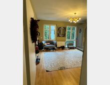 Blick in einen Wohnraum mit Holzboden und großem gemustertem Teppich. An der gelb gestrichenen Wand befinden sich Fenster mit Blick ins Grüne, daneben ein Spiegel und eine Sitzbank, unter der Schuhe aufgereiht sind; ein Kronleuchter beleuchtet den Raum.