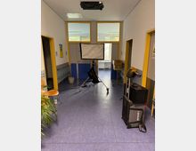 Flurbereich eines Gebäudes mit blauem Linoleumboden und gelb umrahmten Türen und Fenstern. In der Mitte steht ein Lichtstativ mit Softbox, umgeben von technischen Ausrüstungsgegenständen wie einem Koffer und einer Tasche; Zimmerpflanze und Stühle am linken Rand.