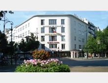 Eckgebäude in modernem Stil mit abgerundeter Fassade, beschriftet mit „Maison de France“, „Institut Français“ und „Cinema Paris“. Im Vordergrund befinden sich eine bepflanzte Verkehrsinsel mit Blumen sowie eine belebte Straßenkreuzung mit Autos, Fahrrädern und Fußgängern.
