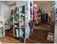 Ein kleines Geschäft mit Holzboden und einer Treppe, die zu einem oberen Bereich führt. Regale und Ständer präsentieren eine Auswahl an Geschenkartikeln, Karten, Dekorationen, Kleidung und Accessoires in einem gemütlichen, hell beleuchteten Raum.