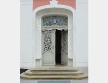 Weiße Doppeltür mit filigranen Ornamenten in einem cremefarbenen Türrahmen, flankiert von weißen, mit Vertikalrillen verzierten Wänden. Oberhalb der Tür befindet sich ein geschwungenes Gitter mit einer integrierten Lampe; vor der Tür führen halbrunde Steinstufen hinauf.