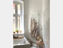 Helles Badezimmer mit einer weißen Badewanne, daneben ein kleines Holzregal mit einem Krug, Zweigen und Handtüchern. An der Wand hängt ein weißer Handtuchheizkörper mit beige Handtüchern, und auf der Fensterbank stehen Blumentöpfe.
