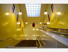 Innenansicht einer Treppenanlage in einer U-Bahn-Station mit gelb gefliesten Wänden, roten Fenstereinfassungen und mehreren Deckenleuchten. Rechts führt eine Rolltreppe nach oben, auf der eine Person zu sehen ist, und über der Szene ist ein großes Oberlichtpaneel.