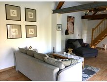 Wohnzimmer in einem modernen Landhausstil mit grauem Sofa, Recamiere und Holzschreibtisch auf einem gemusterten Teppich. Die Wände sind mit gerahmten Bildern dekoriert, und eine Holztreppe führt in einen oberen Bereich.