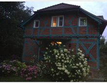 Fachwerkhaus aus Backstein mit türkisfarbenen Holzbalken, umgeben von blühenden Sträuchern, darunter weiße und rosa Hortensien, in abendlicher Stimmung. Im Hintergrund sind Bäume sichtbar, und einige Fenster des Hauses sind beleuchtet.