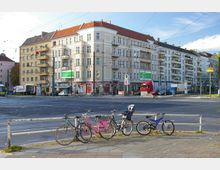 Straßenszene in einer Stadt mit einem mehrstöckigen Wohn- und Geschäftshaus im klassischen Stil, Einzelhandelsgeschäften im Erdgeschoss und Balkonen an den oberen Etagen. Im Vordergrund steht ein Geländer, an dem mehrere Fahrräder angelehnt sind.
