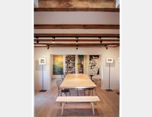 Großer, minimalistischer Raum mit einem langen Holztisch in der Mitte, umgeben von Stühlen und einer Holzbank. Die Decke zeigt sichtbare Holzbalken, während die Wände mit großen Kunstwerken und eingebauten Bücherregalen dekoriert sind; Stehlampen flankieren die Kunstwerke.