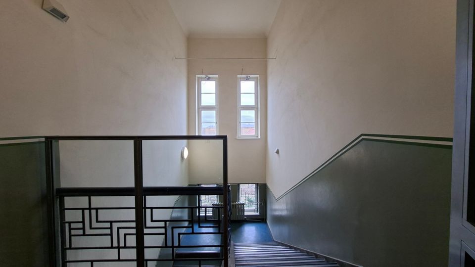 Treppenhaus in einem Mehrfamilienhaus mit hohen, hellen Wänden und einem dunkelgrünen Wandstreifen entlang der Treppe. Ein Metallgeländer mit geometrischem Muster führt hinab zu zwei hohen Fenstern am Treppenabsatz, durch die Tageslicht einfällt.