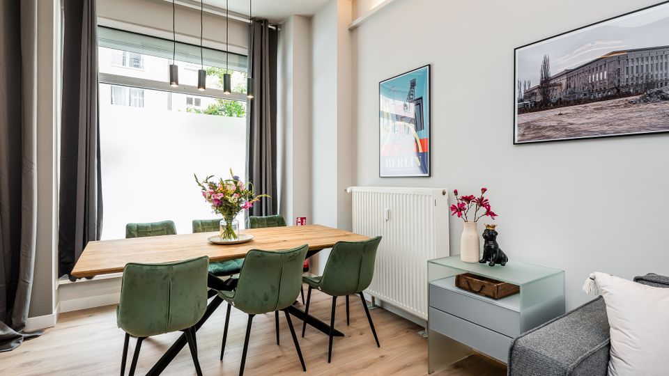 Heller, modern eingerichteter Essbereich mit großem Holztisch und vier grünen Polsterstühlen vor einem bodentiefen Fenster mit dunklen Vorhängen und drei Pendelleuchten. An der weißen Wand hängen gerahmte Bilder; neben einem Heizkörper steht ein kleines Sideboard mit Dekoration, rechts ist ein Teil eines grauen Sofas sichtbar.