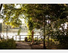 Ein schattiger Pfad führt durch einen Baum- und Buschbestand zu einem Uferbereich mit einem kleinen Holzzaun. Im Hintergrund ist ein ruhiger See zu sehen, umgeben von Bäumen, und bunte Wimpel hängen an einer Schnur.