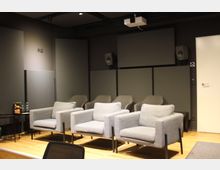 Ein moderner, kleiner Besprechungsraum mit grauen gepolsterten Sesseln in zwei Reihen, einem Holzboden und schallisolierenden Paneelen an den Wänden. Im Raum befindet sich ein Deckenprojektor sowie Lautsprecher, und an der Seite ist ein kleiner Tisch mit Getränken.