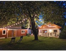 Ein gemütlicher Gartenbereich mit grünem Rasen bei Abenddämmerung, beleuchtet von Lichterketten, die zwischen einem großen Baum und einer kleinen Holzhütte mit überdachter Terrasse gespannt sind. Auf der Terrasse stehen Tische und Stühle, während vor der Hütte zwei Klappstühle aufgestellt sind.