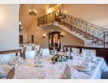 Ein festlich gedeckter Speisesaal mit hellen Wänden und geschwungener Treppe aus Stein mit verziertem Geländer. Die weißen Tische sind mit Geschirr, Gläsern, Kerzenleuchtern und Blumendekoration arrangiert, und im Hintergrund hängen Kronleuchter von der Decke.