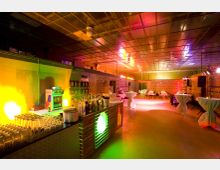 Industrieller Veranstaltungsraum mit einer Bar im Vordergrund, dekoriert mit Flaschen und Gläsern, und runden Stehtischen mit weißen Tüchern. Die Umgebung ist von farbiger Beleuchtung in Gelb, Rot und Violett geprägt, mit einer metallverkleideten Decke und einer Rückwand, die aus Schalttafeln besteht.