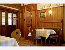 Holzvertäfeltes Interieur eines traditionellen Restaurants mit kariert gedeckten Tischen, Holzstühlen und einer Holzbänken. Eine Tür mit Glasfenstern und dekorative Elemente wie Gemälde und Wandlampen sind sichtbar.