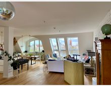 Wohnzimmer mit offener Raumgestaltung und Dachschrägen, großen Fenstern mit Aussicht auf eine städtische Skyline, Holzboden, moderner Einrichtung und einer Mischung aus Sitz- und Essbereich. Im Vordergrund eine Discokugel und Zimmerpflanzen, im Hintergrund ein alter Holzschrank und dekorative Beleuchtungselemente.