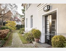 Eingang eines weißen Wohnhauses mit modernen schwarzen Türen und Fenstern, umgeben von einem gepflasterten Weg, gepflegten Sträuchern und herbstlichem Laub. Im Hintergrund ist ein weiteres Haus mit rotem Dach zu sehen.