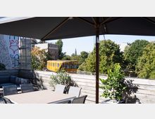 Rooftop-Terrasse mit modernem Sitzbereich und Pflanzen im Vordergrund, im Hintergrund eine gelbe U-Bahn, umgeben von Gebäuden, graffitibemalter Wand und grünen Bäumen. Im Abstand ist eine Kirchturmspitze sichtbar.