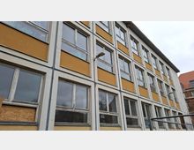 Schräg aufgenommene Fassade eines mehrstöckigen Gebäudes mit gelb-weißen Wandfeldern und vielen rechteckigen Fenstern in gleichmäßigen Rasterreihen. An der Außenwand sind eine Lampe und teils beschädigte bzw. verschlossene Fensterbereiche zu sehen; im Hintergrund ragen Dächer und ein bewölkter Himmel auf.