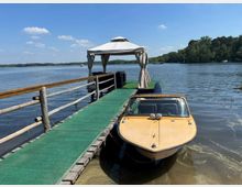 Holzsteg mit grünem Bodenbelag führt über einen ruhigen See zu einem kleinen Pavillon mit weißem Dach und Vorhängen. Rechts liegt ein Motorboot am Steg, im Hintergrund sind bewaldete Ufer und blauer Himmel zu sehen.