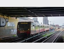Eine S-Bahn fährt unter einer Brücke auf einem mehrgleisigen Bahngleis, mit Graffiti an der Brückenwand. Im Hintergrund sind moderne Hochhäuser, Baukräne und weitere Bahngleise zu sehen.