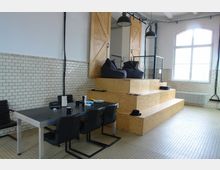 Industrie-inspirierter Innenraum mit weiß gefliesten Wänden, großen Fenstern und schwarzem Metallrahmen. Im Vordergrund steht ein Tisch mit schwarzen Stühlen, daneben eine mehrstufige Sitzplattform aus hellem Holz mit Sitzsäcken und Hängelampen an der Decke.