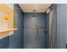 Geflieste Duschkabine mit blauen Wandfliesen, Regenduschkopf an der Decke und Handbrause mit gelben Armaturen an der Rückwand. Rechts hängt ein grauer Duschvorhang an einer Stange, links ist ein gelbes Drahtregal mit Ablage zu sehen.