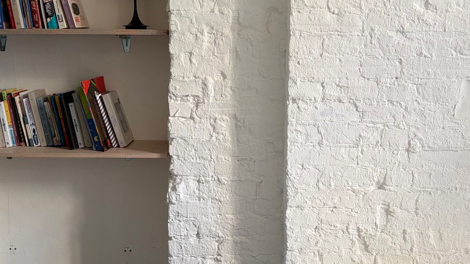 Minimalistischer Innenraum mit weiß gestrichener Backsteinwand und einem eingebauten Regal mit Büchern und kleiner Tischlampe. Daneben stehen ein blauer Hocker auf Betonboden und ein Bett mit weißer Bettwäsche am rechten Bildrand.