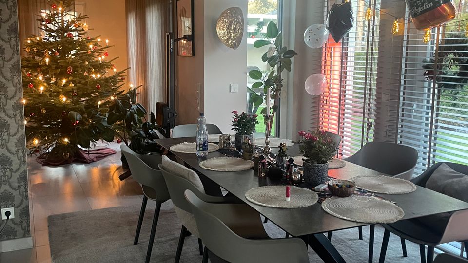 Ein modern eingerichtetes Esszimmer mit einem langen Esstisch, umgeben von mehreren Stühlen. Im Hintergrund steht ein geschmückter Weihnachtsbaum mit Lichtern, rechts sind große Fenster mit Lamellenjalousien und Blick auf einen Garten.