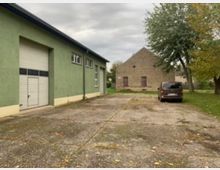 Grünes Lagergebäude mit weißem Rolltor links und einem steinernen, fensterlosen Gebäude mit zwei Holztüren im Hintergrund. Davor eine asphaltierte Fläche mit Laub und ein geparktes Auto auf der rechten Seite, umgeben von Bäumen und Grünflächen.