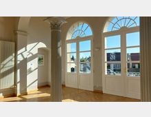 Helle Innenansicht eines Raumes mit großen, weißen Doppeltüren und halbkreisförmigen Fenstern oben, durch die Licht einfällt. Im Vordergrund ist eine verzierte Säule erkennbar, während der Boden aus Holzparkett besteht und an der linken Wand ein Heizkörper angebracht ist.