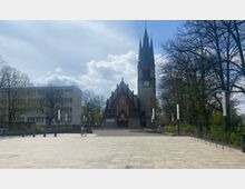 Kirchengebäude mit einem hohen Turm und einer Fassade aus roten Ziegeln und Stein, umgeben von kahlen Bäumen und einem gepflasterten Platz; daneben ein modernes, mehrstöckiges Gebäude. Im Vordergrund fährt eine Person auf einem Fahrrad.