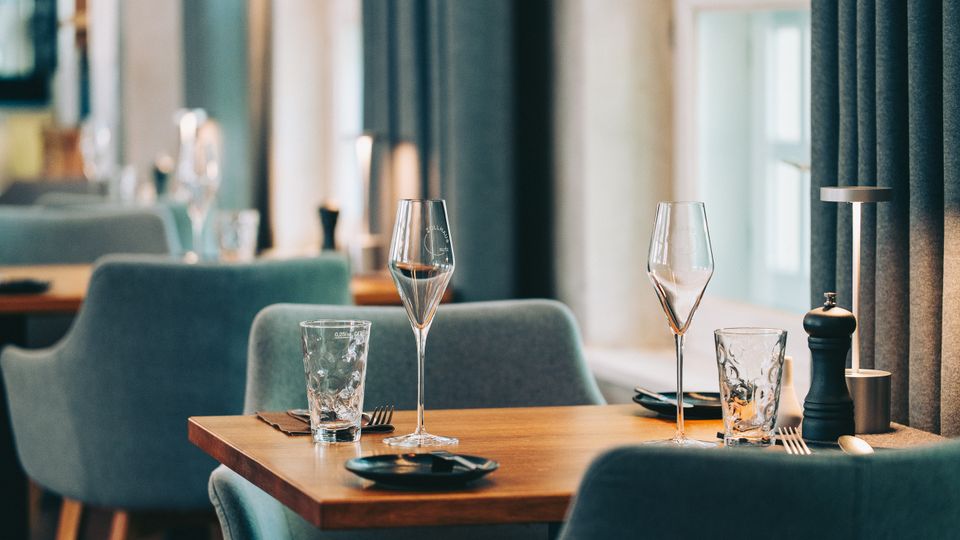 Modernes Restaurantinterieur mit Holztischen und gepolsterten, blaugrauen Stühlen; auf dem vorderen Tisch stehen Wein- und Wassergläser, Besteck, Teller sowie eine Pfeffermühle. Große Fenster mit dunklen Vorhängen lassen Tageslicht in den Raum.