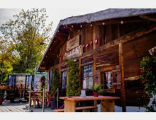 Rustikale Holzhütte mit Satteldach und dunklen Holzfassaden, geschmückt mit Lichterkette und kleinen rot-weißen Wimpeln, vor großen Fenstern mit Herzläden. Davor steht auf einer Holzterrasse ein kleiner Außenbereich mit Holztischen, Bänken, Pflanzen und Absperrkordeln, umgeben von Bäumen und Blumen.