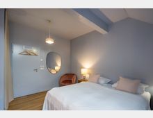 Ein schlichtes Schlafzimmer mit hellblauen Wänden, einem großen Bett mit weißer Bettwäsche und cremefarbenen Kissen. Im Raum befinden sich eine kleine Nachttischlampe, ein runder Spiegel, ein rosa Sessel, und an der Tür hängen Kleiderbügel.