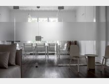 Moderner Besprechungsraum mit Glaswand, die teils blickdicht foliert ist. Im Raum stehen ein weißer Konferenztisch mit mehreren Stühlen, ein Fenster sorgt für Tageslicht, und auf der linken Seite ist eine graue Polsterecke teilweise sichtbar.