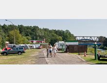 Ein Campingplatz mit parkenden Wohnwagen, Autos und Zelten am Waldrand neben einem Fluss. Im Hintergrund ist eine Eisenbahnbrücke zu sehen, während ein Paar auf einem Weg neben den Fahrzeugen spaziert.