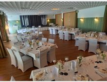 Ein dekorierter Veranstaltungsraum mit festlich eingedeckten Tischen, weißen Tischdecken und Stuhlhussen, Blumenarrangements und Kerzen. Im Hintergrund befindet sich eine kleine Bühne mit Vorhängen und modernem Decken- und Wanddesign.