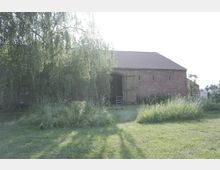 Backsteingebäude mit Satteldach, dessen Holztor geöffnet ist, umgeben von Gras und einer großen hängenden Weide. Im Vordergrund befindet sich eine Grünfläche mit dichtem Bewuchs.