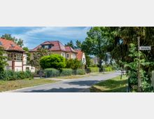 Straßenszene in einem Wohngebiet mit freistehenden Häusern, roten Ziegeldächern und gepflegten Gärten, flankiert von Bäumen und Sträuchern; Straßenschild mit der Aufschrift „Gotensteg“ sichtbar.