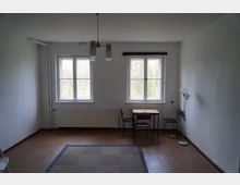 Ein schlicht eingerichteter Raum mit weißen Wänden, zwei Fenstern mit Blick ins Grüne, einem Teppich auf dem braunen Boden und einer einfachen Tisch- und Stuhlkombination in der Ecke. An der Decke hängt eine einfache Lampe, und an der rechten Wand steht eine Stehlampe.