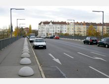 Städtische Straße mit einem Bürgersteig auf der linken Seite, der durch halbkugelförmige Betonbarrieren von der Fahrbahn getrennt ist. Im Hintergrund sind mehrstöckige Wohnhäuser mit weißen und pastellfarbenen Fassaden sichtbar, und am Straßenrand parken Autos.