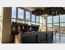 Innenraum eines Gebäudes mit einer Bar aus Holz und Glas. Große Fensterfronten lassen viel Tageslicht herein und bieten einen Blick auf Gebäude und Bäume im Außenbereich. Die Decke ist mit modernen, röhrenförmigen Designelementen dekoriert.