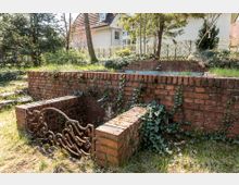 Gartenbereich mit kleinen Backsteinmauern, Efeubewuchs und einem dekorativen schmiedeeisernen Geländer. Im Hintergrund steht ein helles Wohnhaus, umgeben von Bäumen und Sträuchern.