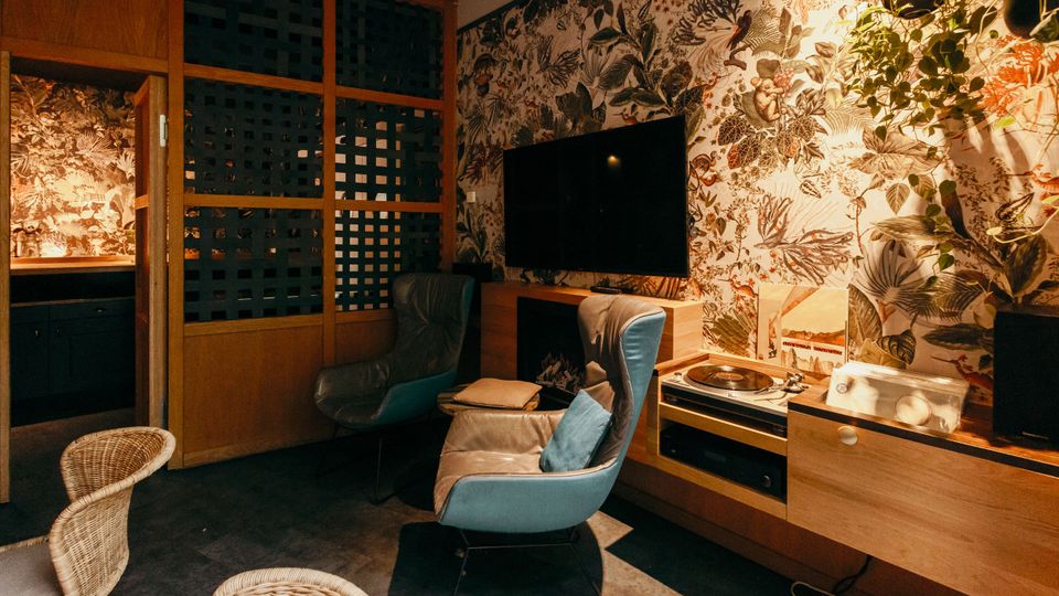 Gemütlicher Lounge- oder Wohnzimmerbereich mit gemusterter Tapete, zwei gepolsterten Sesseln und einem wandmontierten Fernseher über einem Holzsideboard. Rechts stehen ein Plattenspieler und weitere Audiogeräte, links trennt eine Holzschiebetür mit Gitterelementen den Raum von einer angrenzenden Nische.