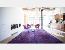 Helles Wohnzimmer in einem Loft mit Backsteindecken, einem violetten Teppich, modernen Stühlen in Rosa und Rot, einer Wand mit Kamin, sowie offener Küche mit Holzverkleidung auf der rechten Seite. Große Fenster im Hintergrund lassen viel Tageslicht herein.