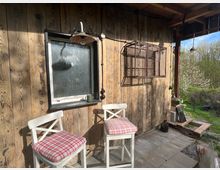 Überdachte Veranda an einer Holzhütte mit Bretterwand, kleinem Fenster, Wandlampen und einem angebrachten Drahtregal. Davor stehen zwei weiße Barhocker mit karierten Sitzkissen auf Steinplatten; rechts öffnet sich der Blick ins grüne Gelände.