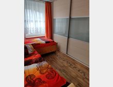 Schlafzimmer mit zwei Einzelbetten aus Holz, bunten Bettdecken in Rot- und Orange-Tönen, einem großen Kleiderschrank mit Schiebetüren in Holzoptik und Milchglas, sowie einem Fenster mit weißen Spitzenvorhängen und orangefarbenem Verdunkelungsvorhang. Der Boden ist mit Holzfliesen und einem dekorativen Muster gestaltet.