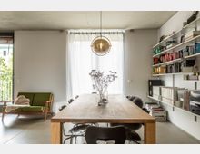 Moderner Wohnraum mit einem großen Esstisch aus Holz, umgeben von schwarzen Stühlen. Im Hintergrund ein großes Fenster mit weißen Vorhängen, rechts Wandregale mit Büchern und Ordnern, links ein grünes Sofa.